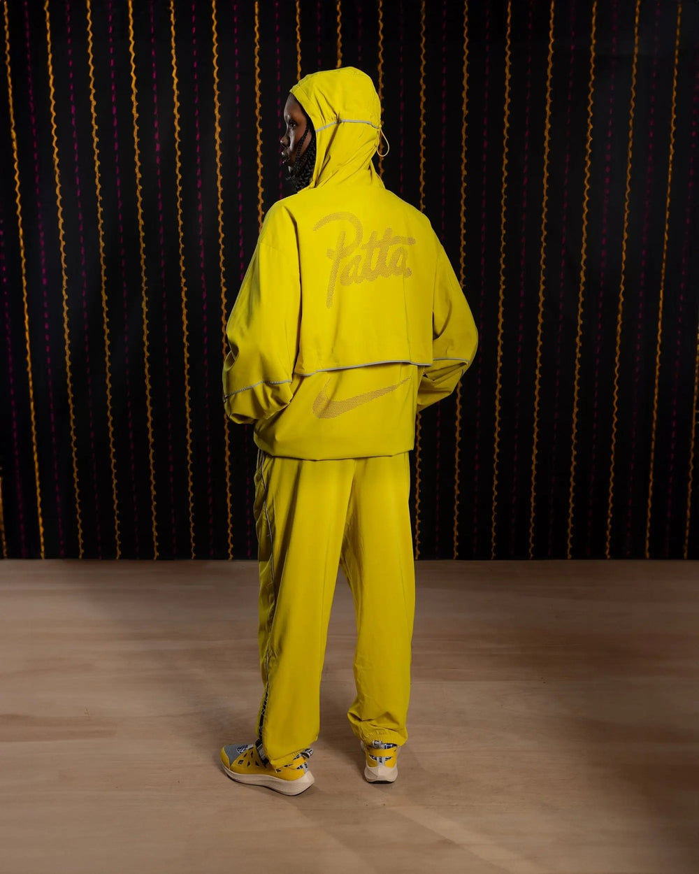 Back view of a model wearing the yellow Nike x Patta Running Team hooded track jacket with logo details.