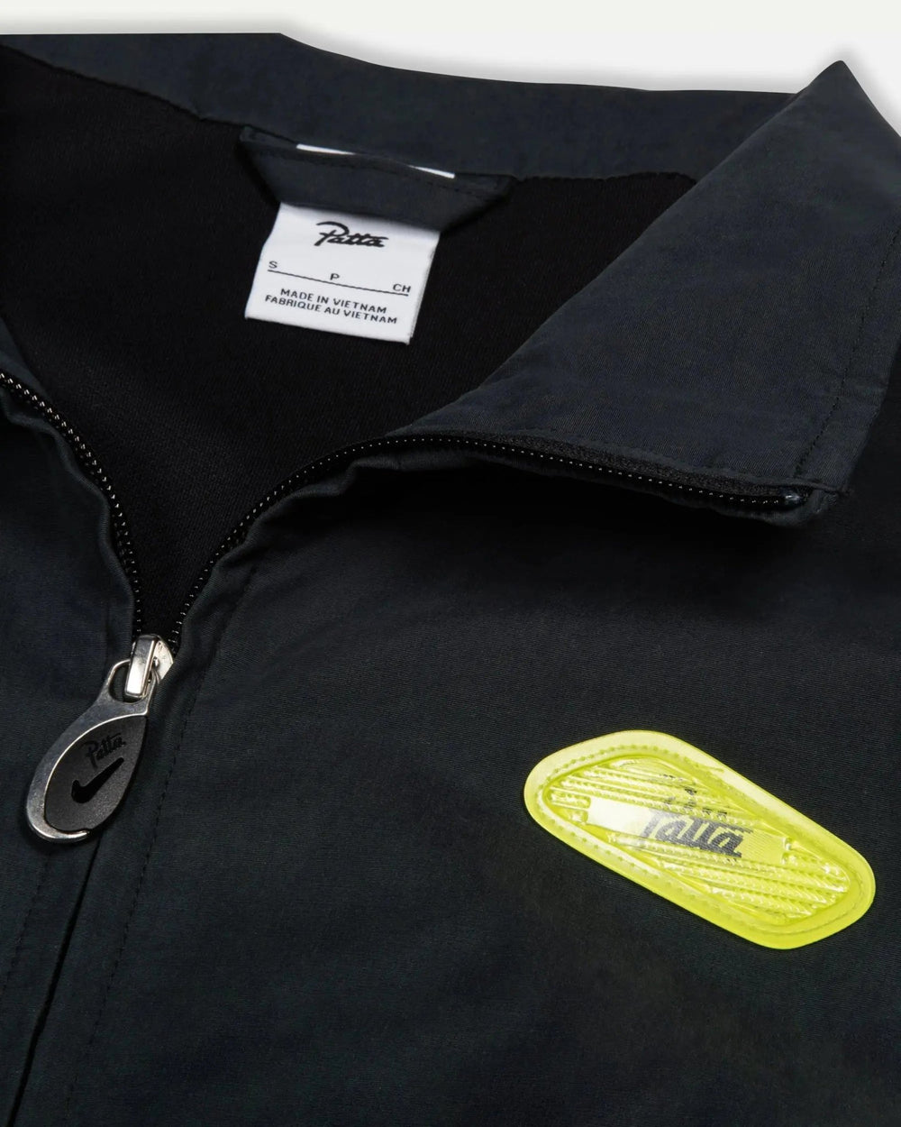 Detail of the Patta x Nike NRG Track Jacket in black, featuring the collar, zipper pull, and cyber yellow badge.